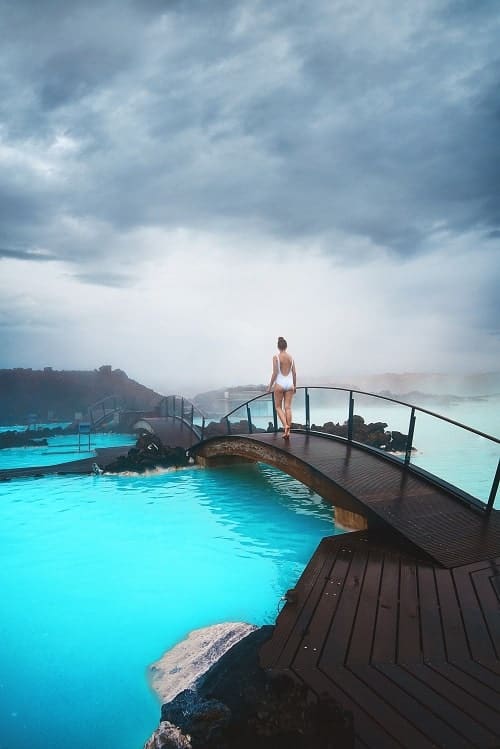 a single person standing on a bridge over a bright blue geothermal pool at the blue lagoon in iceland 
