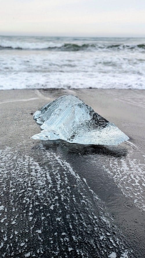a chunk of transparent glacier sitting on a the black sand diamond beach in iceland