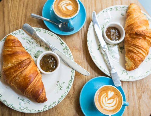 two croissants sitting on china plates next to lattes on a wood table