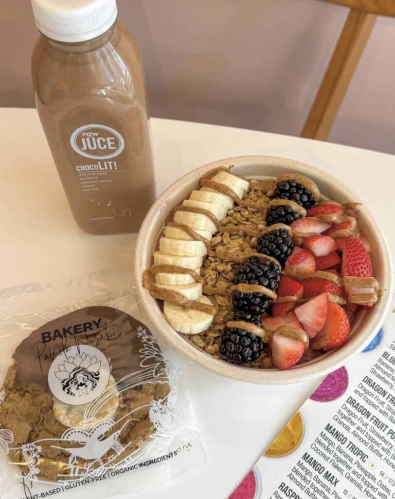 Acai bowl with bananas, blackberries, strawberries and granola next to almond chocolate milk and cookie on a white table.