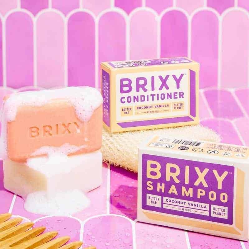 boxes of vegan shampoo bars created by the brand brixy next to a bar covered in suds on a pink tile background