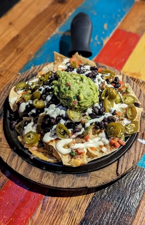 a large skillet of vegan nachos topped with guac, sour cream, and beans sitting on a colorful wood table at nacho daddy in las vegas