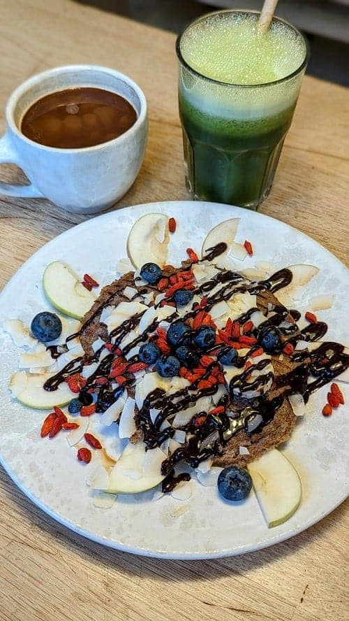 raw vegan pancakes topped with apple slices, chocolate drizzle and goji berrie next to a coffee and green smoothie at 42 raw in copenhagen