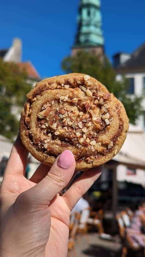 a round vegan cinnamon roll covered in nuts held in front of a church with a copper spire in copenhagen