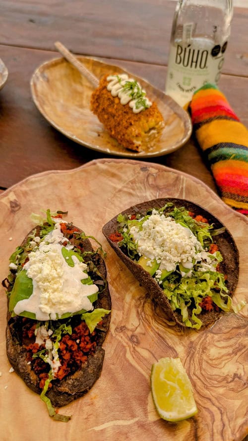 two blue corn tacos with vegan cream sauce and avocado next to a small vegan elote corn