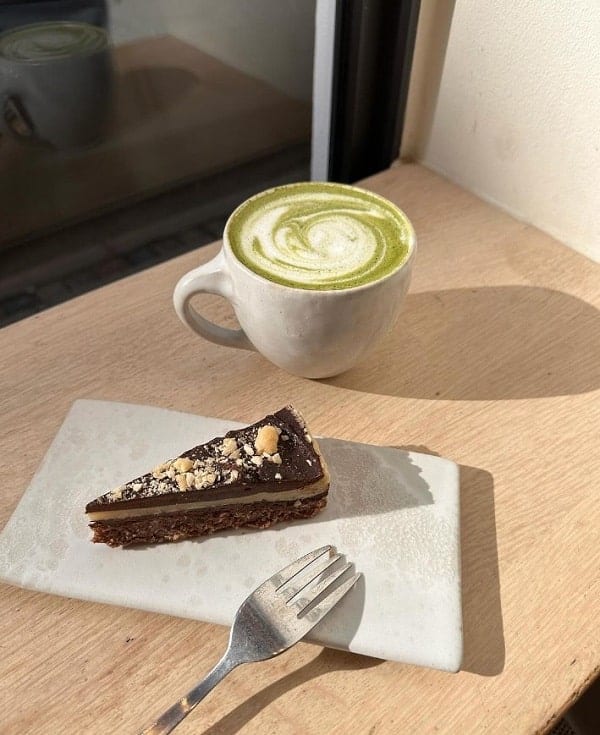 a slice of vegan chocolate cake on a white plate next to a matcha latte on a wood table at 42 raw in copenhagen