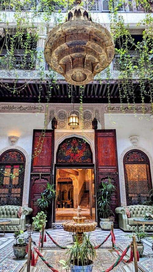 the inside of an ornate riah with dark wood doors, hanging green plants, and a fountain in morocco