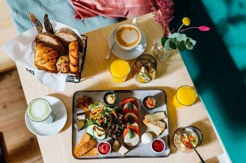 a large vegan brunch tray with pastries, fresh fruit, and veggies next to a coffee and juice on a light wood table at ima vegan in hamburg