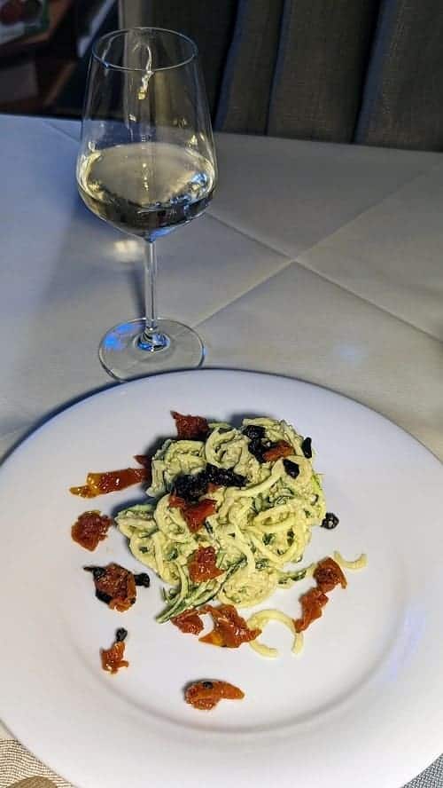 a white plate with a mound of zucchini noodles covered in a cashew cream sauce next to a glass of white wine in venice italy