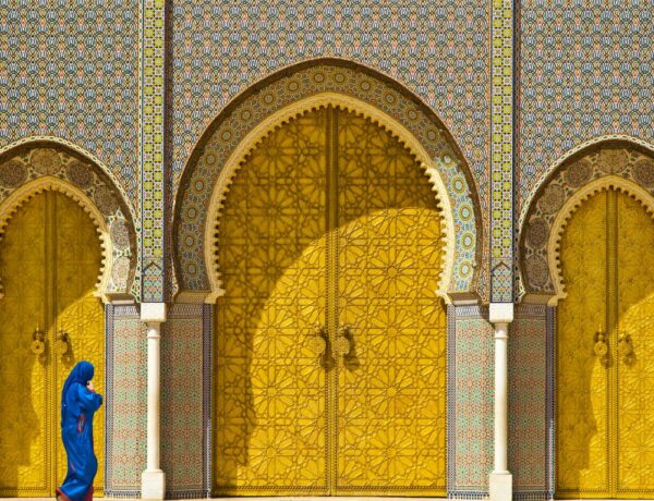 ornate bronze doors surrounded by intricate colorful tile mosaics with a woman walking in front of the door dressed in a full blue dress and head covering in morocco