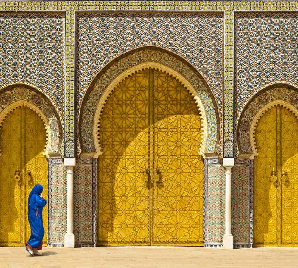 ornate bronze doors surrounded by intricate colorful tile mosaics with a woman walking in front of the door dressed in a full blue dress and head covering in morocco