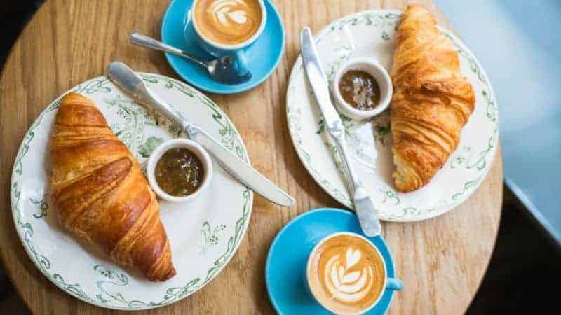 two golden vegan croissants on a wood table next to two coffees