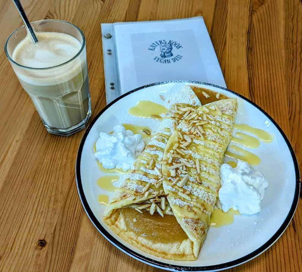 vegan german style pancake filled with apples and covered in whipped cream next to a coffe on a wood table