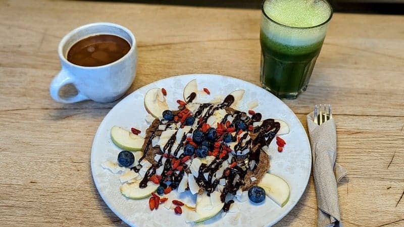 raw vegan pancakes topped with apple slices, chocolate drizzle and goji berrie next to a coffee and green smoothie at 42 raw in copenhagen