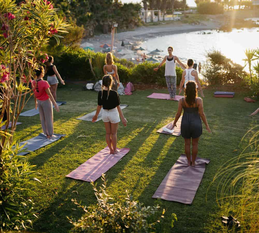 six people with their back to the camera overlooking a beach and lush jungle area doing yoga at sunset