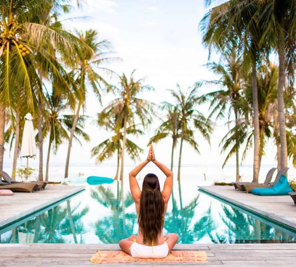 a woman meditating in front of a pool surrounded by palm trees at a wellness retreat