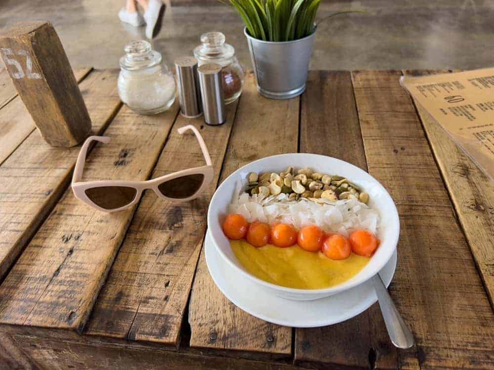 colorful vegan smoothie bowl topped with fruit and coconut sitting on a wood table set to tan sunglasses in bali