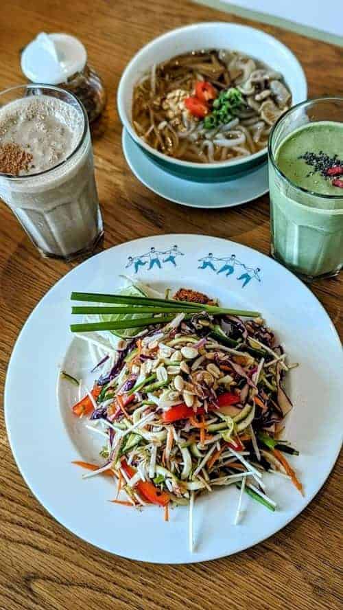 a white bowl with vegan pad thai and a bowl of spicy tom yum soup next to a green and brown smoothie on a wood table