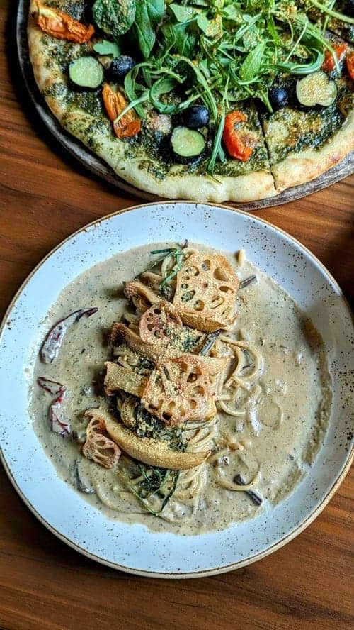 vegan mushroom cream pasta next to a golden pesto pizza on a wood table at lazy farmers