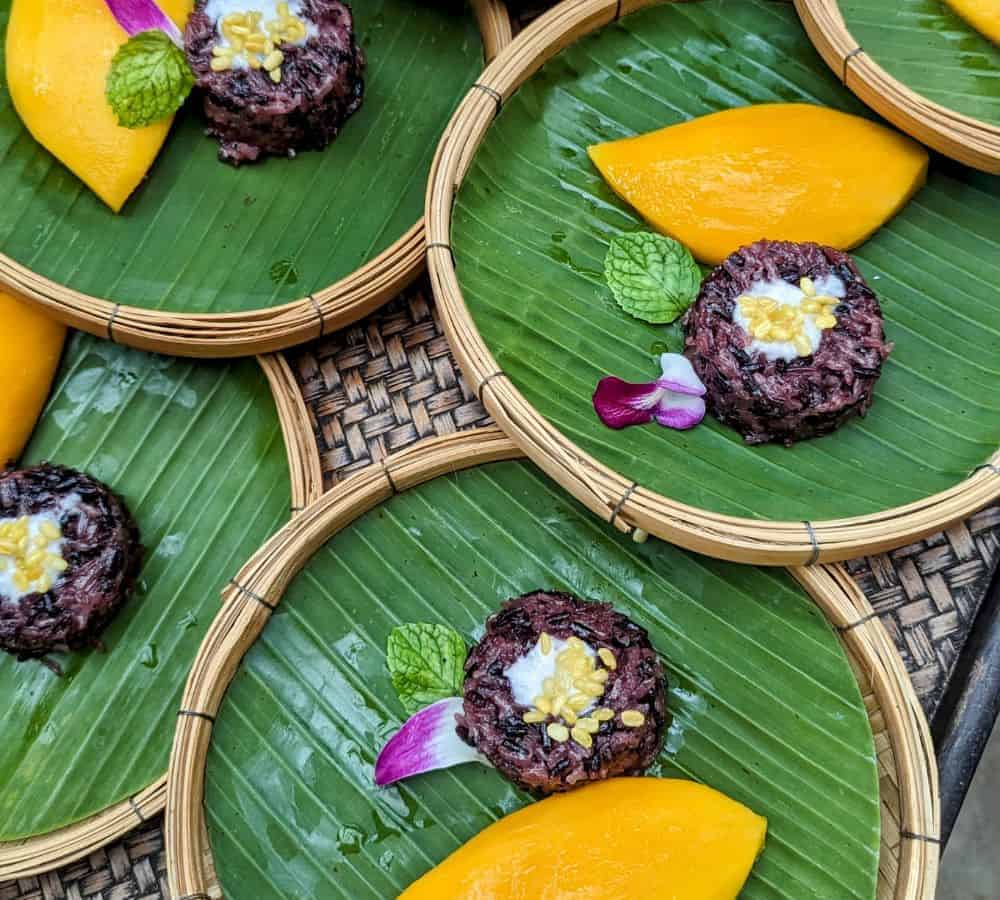small round wooden plates covered with a banana leaf with a hunk of mango and purple coconut rice