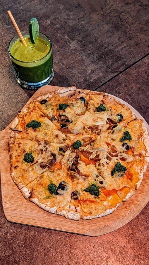 a vegan and gluten free pizza on a wood platter next to a green juice with a lime at the vegan table in bangkok