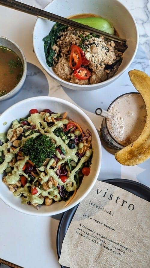 two round white bowls filled with a green kale superfood salad and a noodle dish next to a latte topped with a banana at vegan vistro in bangkok