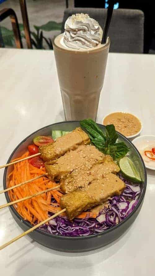 a colorful vegan tofu peanut satay bowl on a white table next to a large peanut butter milkshake topped with vegan whipped cream in bangkok