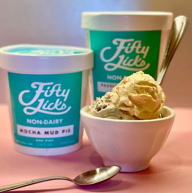 two white and teal pints of vegan ice cream standing next to a white bowl filled with two scoops and a spoon from fifty licks