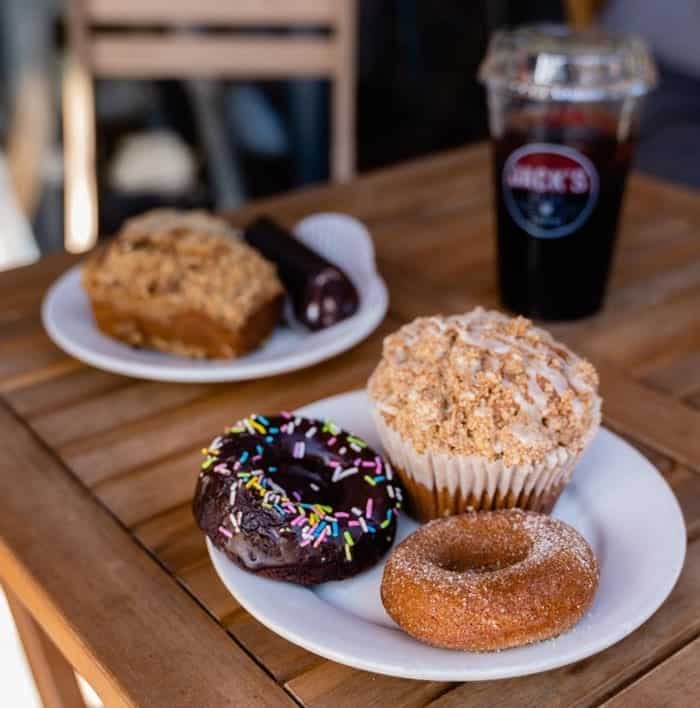 a small round white plate with a plain vegan donut next to a chocolate vegan donut and a muffin with an iced coffee in the background at jack stir's in nyc