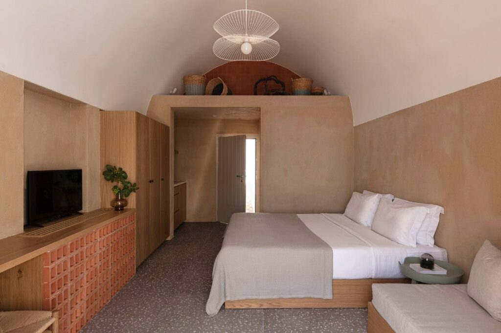 inside of a dimly lit guestroom with neutral walls and a white double bed at ethos vegan hotel in santorini greece