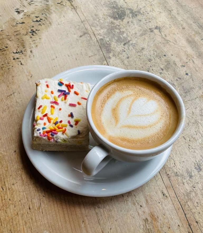 a vegan latte sitting on a plate next to a slice of cake topped with a white buttercream and colorful sprinkles at grindcore house in philly