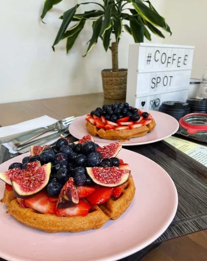 two plates of vegan belgian waffles covered in fresh fruit and slices of figs on a wood table with a coffee sign in the background at heart garden waffles in amsterdam