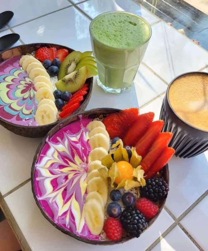 two colorful smoothie bowls topped with a zig zag fruit puree pattern and fresh cut fruit next to a green juice and a coffee at rainbowls in amsterdam