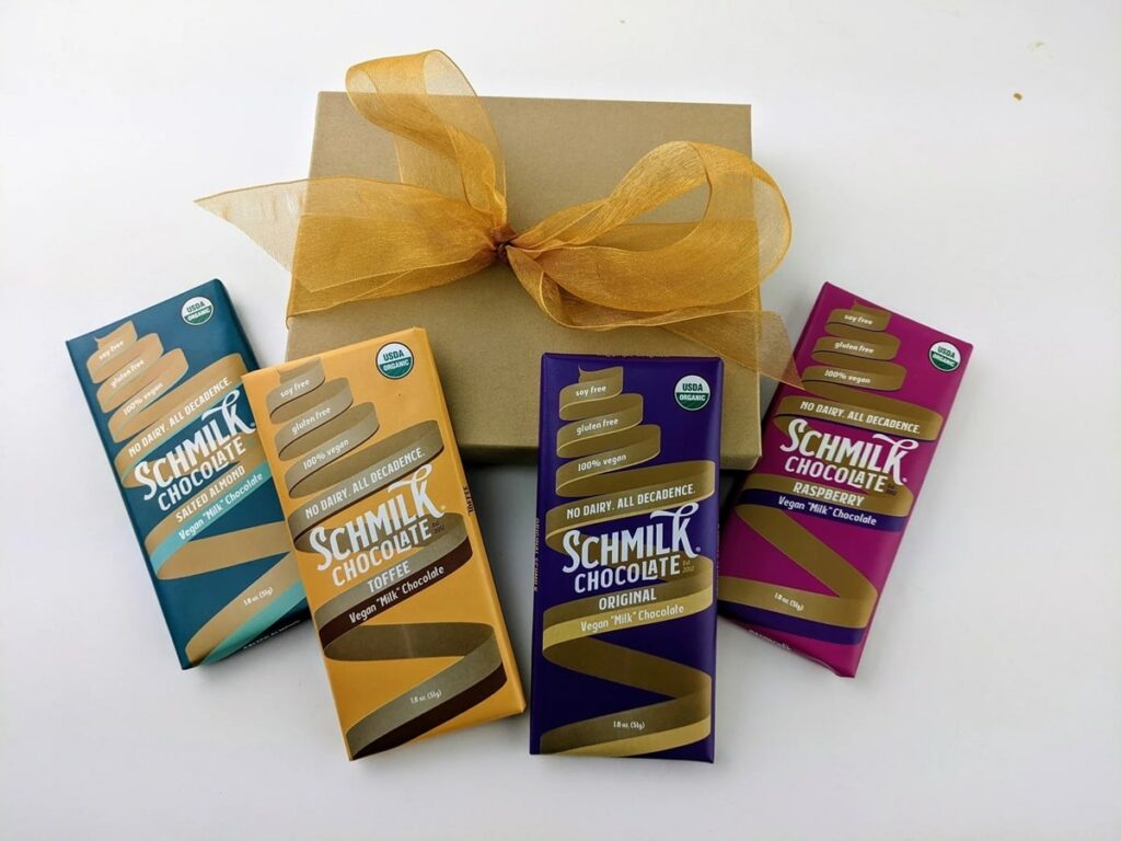 a variety pack of four different vegan milk chocolate bars leaning against a brown box from schmilk