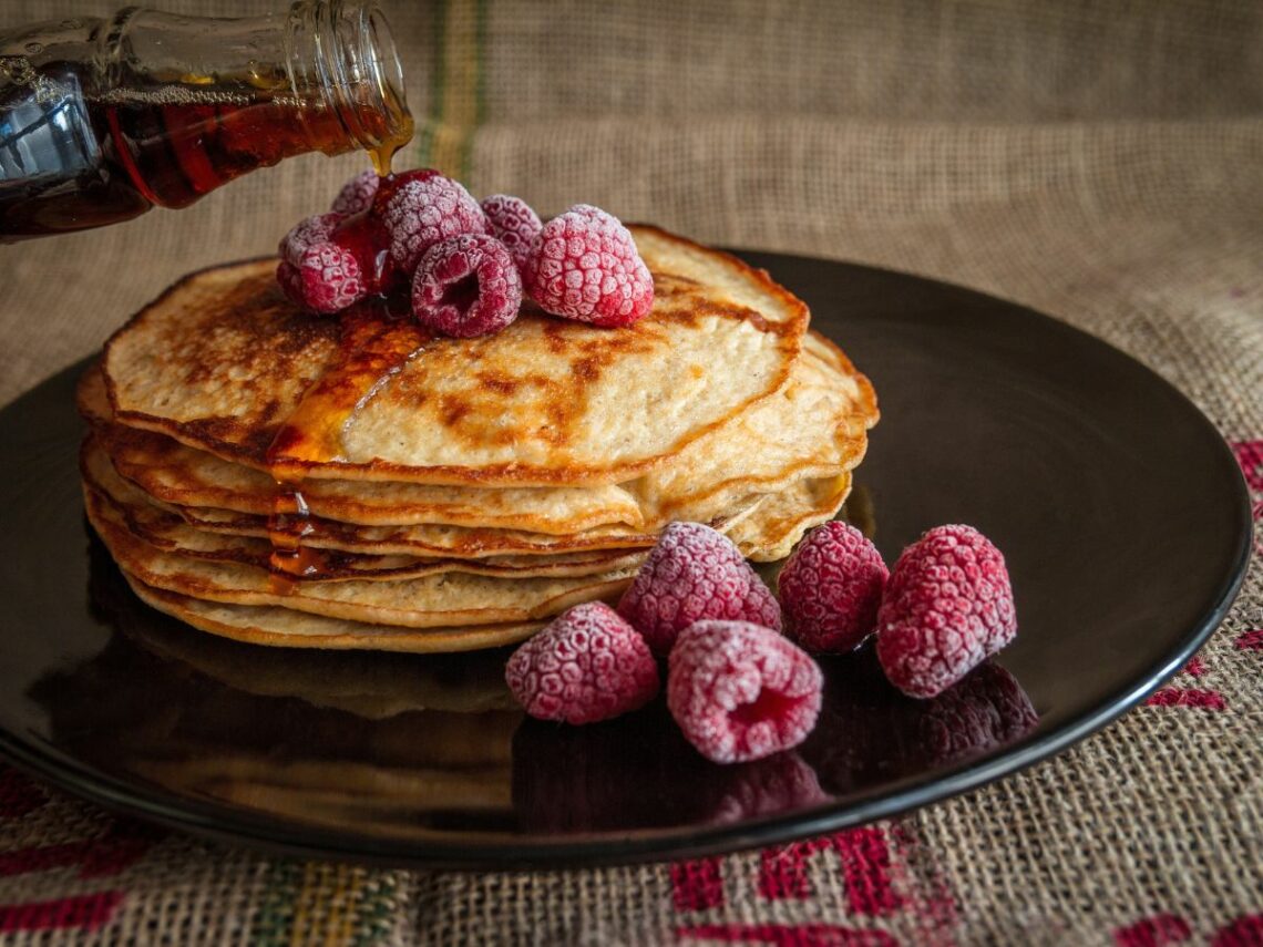 a stack of three golden vegan pancakes being covered in maple syrup and topped with lightly frosted raspberries