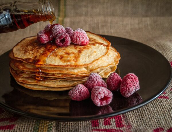 a stack of three golden vegan pancakes being covered in maple syrup and topped with lightly frosted raspberries