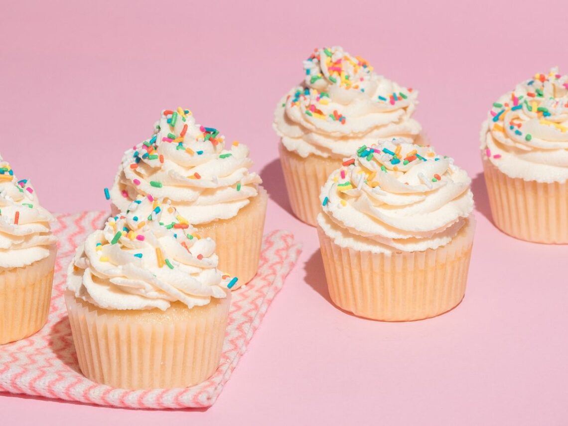 two lines of vegan vanilla cupcakes topped with swirls of white butter cream and topped with colorful sprinkles on a pink background