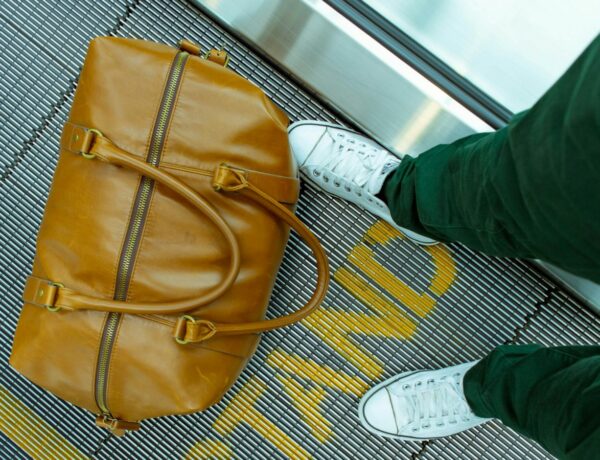 a light tan weekender bag sitting on a silver moving walkway at an airport near someones feet