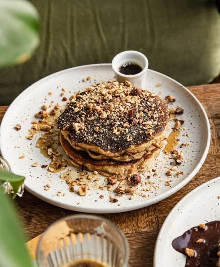 a stack of vegan pancakes sitting on a white plate and topped with a nut crumble with maple syrup at Aujourd’hui Demain in paris