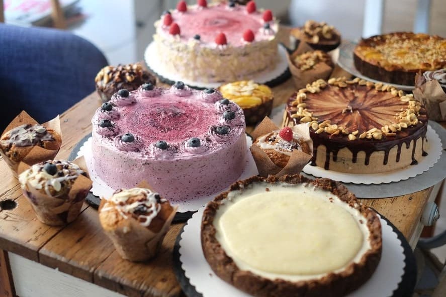 a wood table covered with beautiful vegan whole cakes, pies, and muffins in vienna