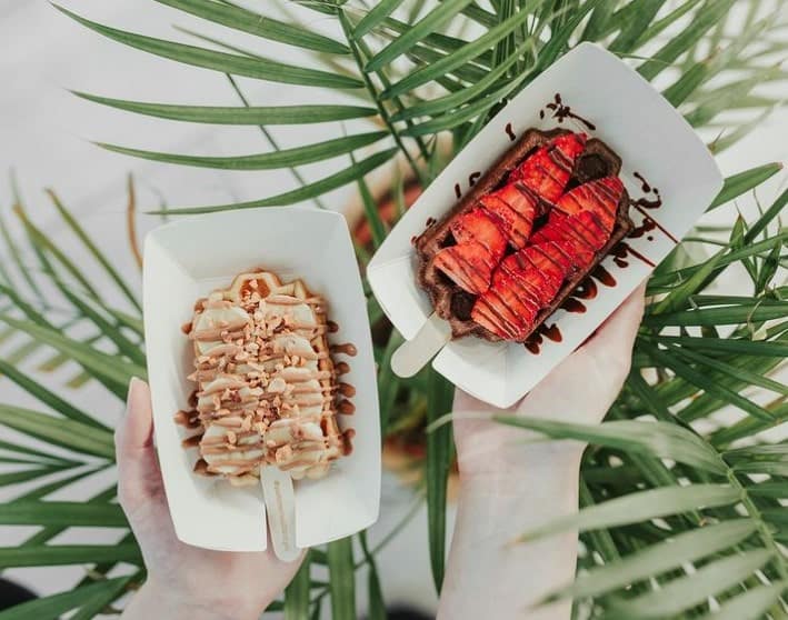 two colorful vegan popsicles in paper trays topped with chocolate, coconut and sliced strawberry in budapest
