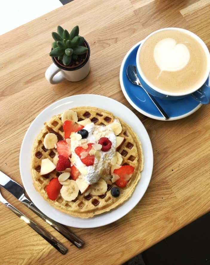a round vegan belgian waffle on a white plate, topped with sliced berries and coconut whipped cream next to a latte in a blue mug at cloud cakes in paris