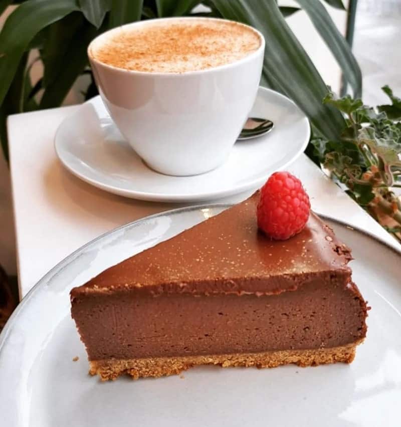 a single slice of dark chocolate vegan cheesecake with a single raspberry on top next to a white mug of coffee with milk foam and a dusting of cinnamon in edinburgh