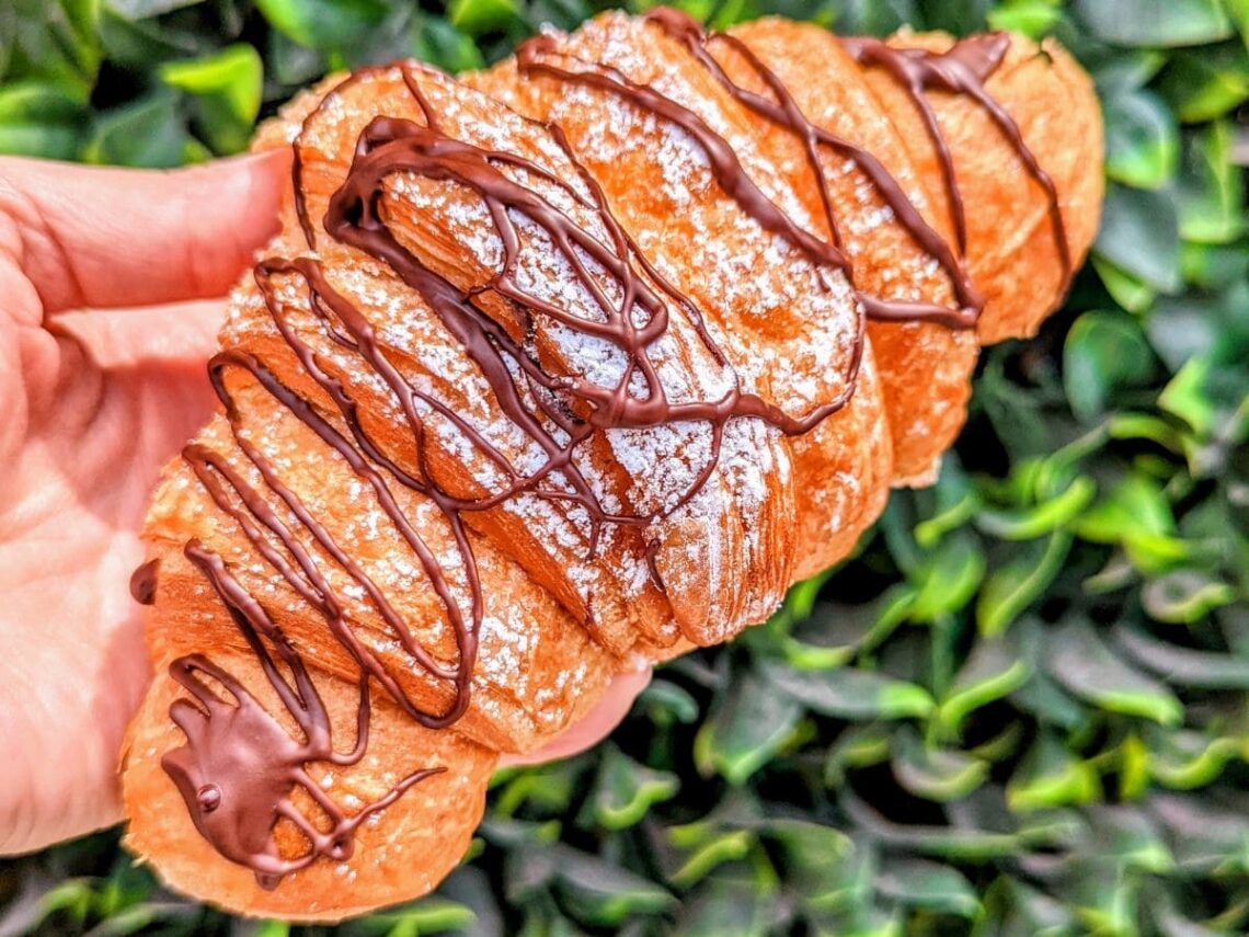 a golden vegan croissant drizzled with chocolate held in front a green leaves