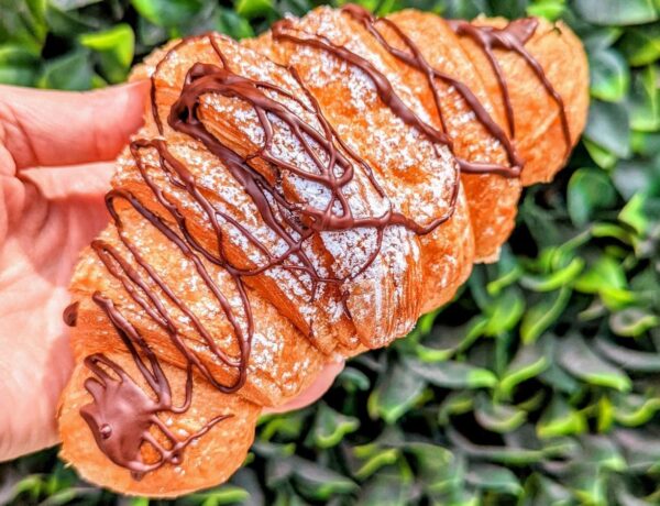 a golden vegan croissant drizzled with chocolate held in front a green leaves