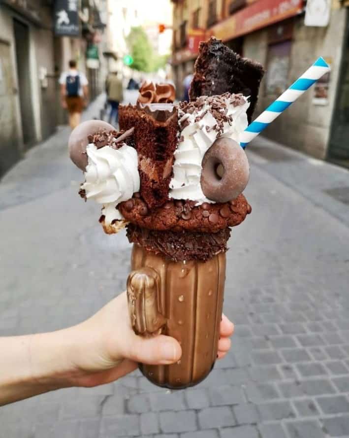 a giant vegan milkshare topped with a donut, brownie, whipped cream and other dessert treats held in the middle of a street in madrid