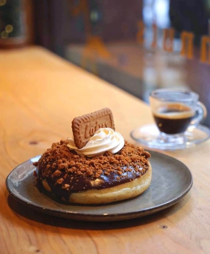 a vegan Lotus donut topped with chocolate, cream, and a lotus cookie sitting on a dark plate next to an espresso shot in madrid