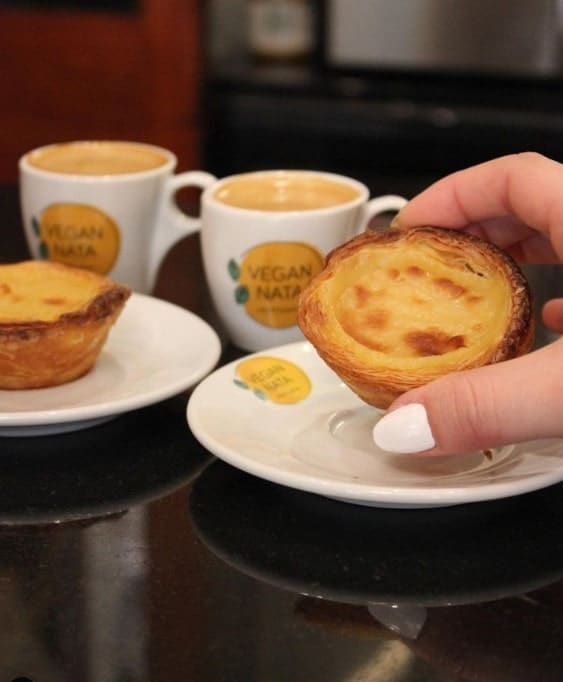 a person holding a golden vegan pastel de nata in front of cups of espresso in lisbon