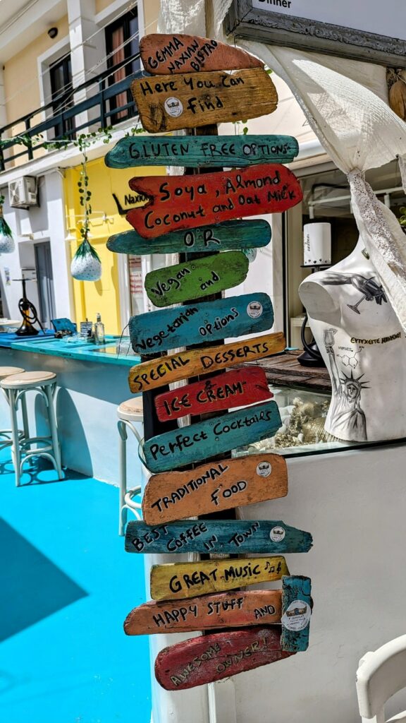 a colorful, worn wooden sign that says vegan, vegetarian, gluten free and more at gemma naxian  in naxos