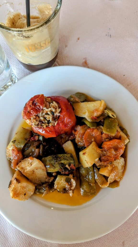 a stuffed greek tomato with roasted veggies in a tomato sauce on a white plate next to an iced coffee